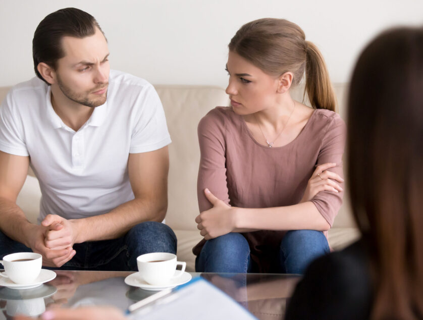 Young angry divorcing couple sitting on couch with arms crossed looking at each other with unconcealed hatred and hate, psychological help with problems in unhappy marriage, family counseling